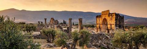 Les ruines romaines de Volubilis à l'heure dorée : arcs de triomphe et colonnes dressées avec les montagnes du Moyen Atlas en arrière-plan