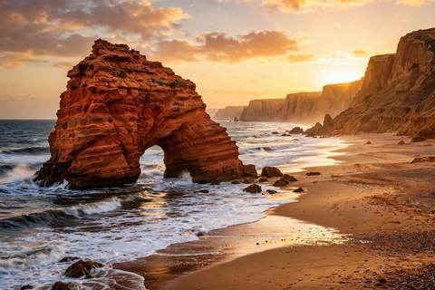 Une immense arche de grès rouge sur la plage de Legzira, au Maroc, avec les vagues de l'Atlantique à sa base et des falaises ocres le long du rivage désert à l'heure dorée